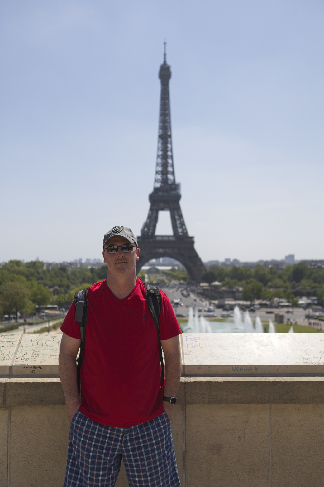 A photograph of a tourist posing in an outdoor setting, with the iconic Eiffel Tower dominating the background under a clear blue sky.