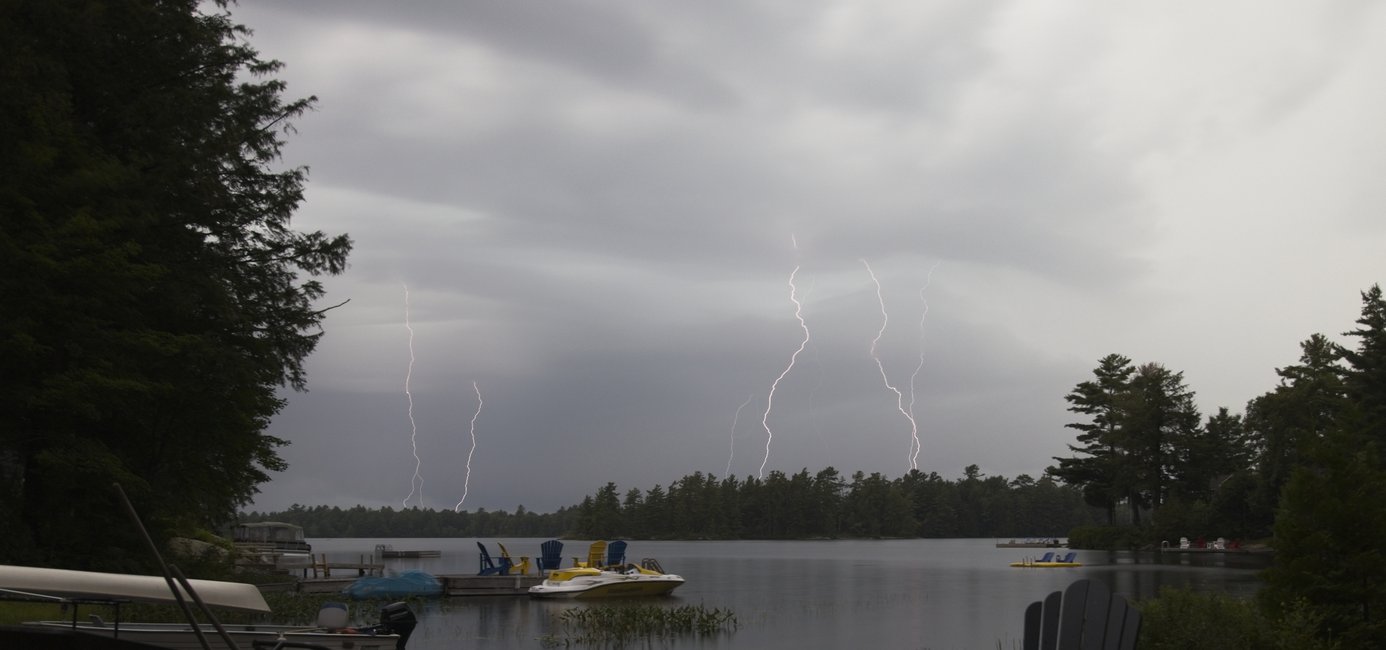 A dramatic lake scene during a thunderstorm, with multiple lightning bolts striking through dark, overcast clouds above a calm body of water. Trees line both shores and a forested ridge stretches across the far side, while a wooden dock in the foreground holds colorful paddle boats, a yellow and white motorboat, and Adirondack chairs. The stormy gray sky contrasts with the peaceful shoreline setting.
