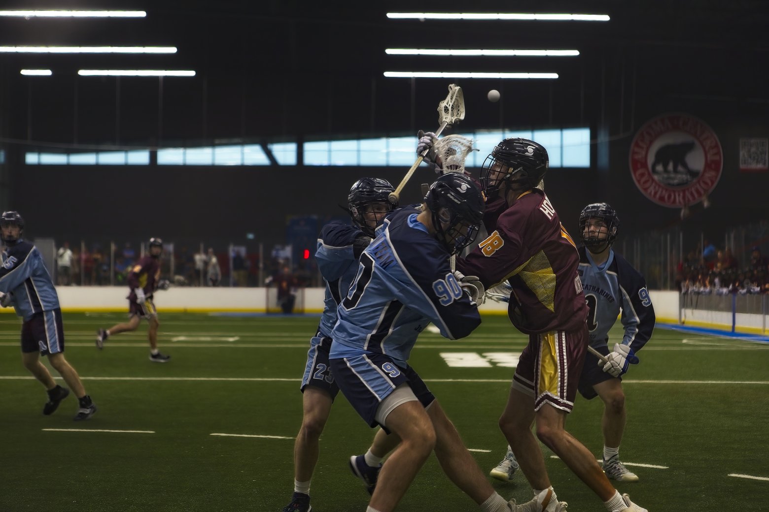 Indoor lacrosse game in progress with players from two teams competing on a green turf field. A player in a light blue jersey (number 90) battles for position against a player in a maroon jersey (number 88) while the lacrosse ball is airborne near the nets. Additional players in both light blue and maroon uniforms are positioned around the field, and a 'VICTORIA CENTRAL CORPORATION' sign with a bear logo is visible in the background.